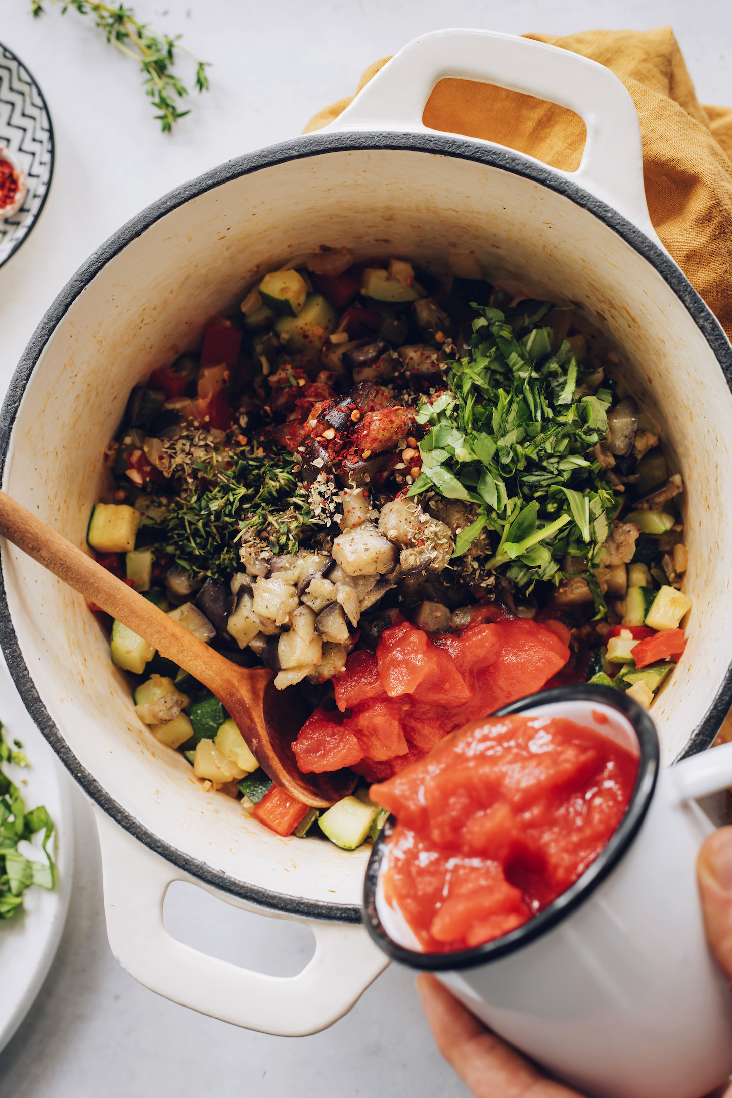 Pouring canned tomatoes over sautéed eggplant, zucchini, and bell pepper with fresh herbs