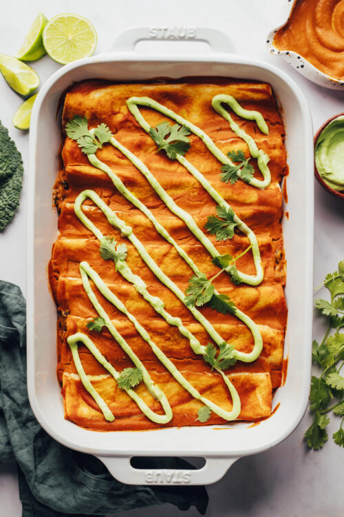 Ceramic baking dish filled with pumpkin black bean enchiladas drizzled with avocado crema