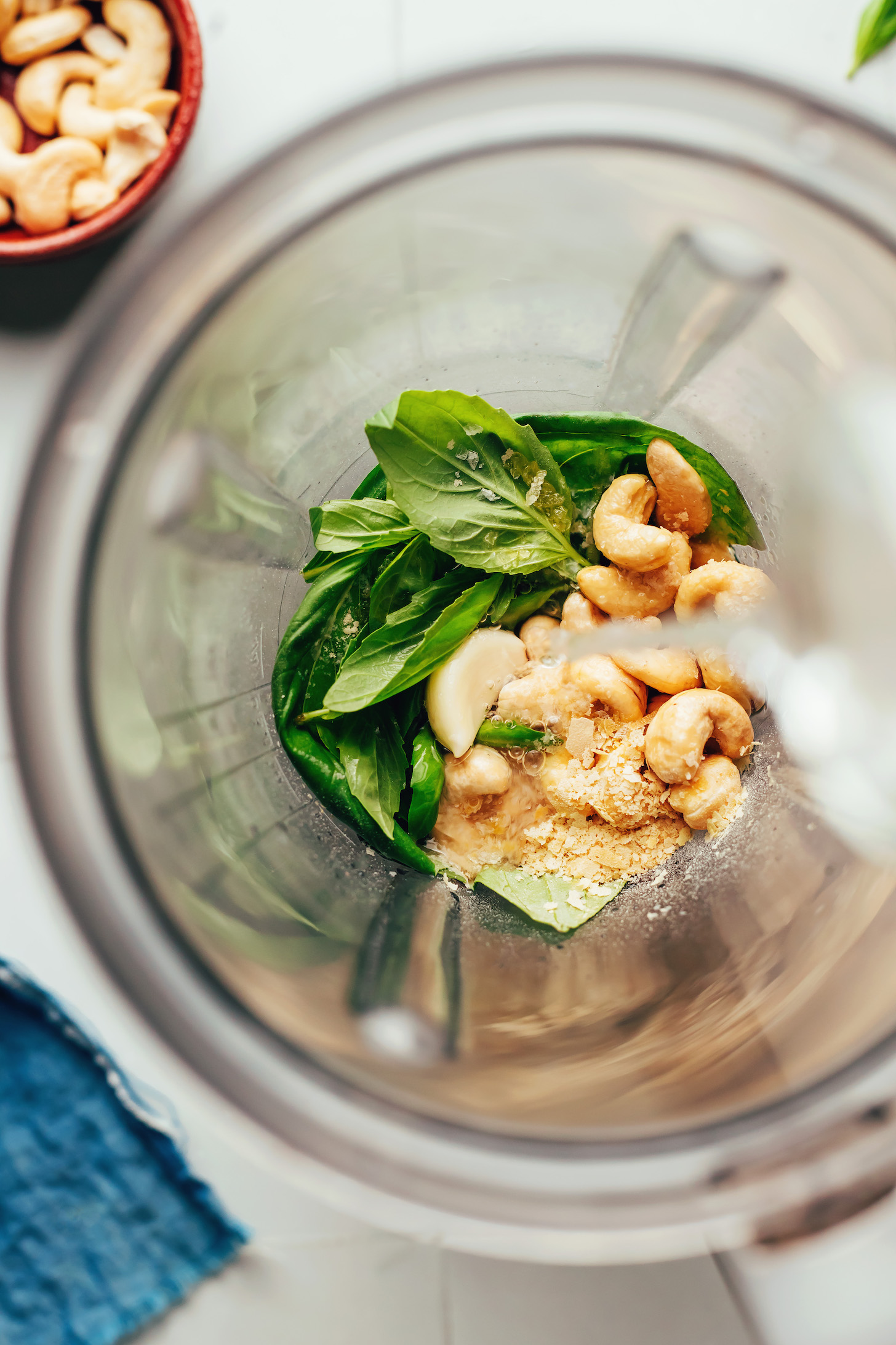 Pouring water into a blender over cashews, garlic, fresh basil, and nutritional yeast