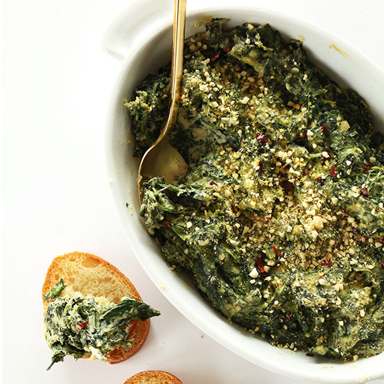 Slice of bread topped with Creamy Kale and Spinach Dip with the rest of the pan of dip beside it