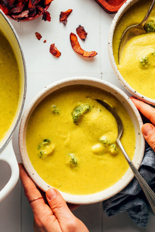 Overhead shot of hands holding the sides of a bowl of creamy vegan broccoli cheddar soup