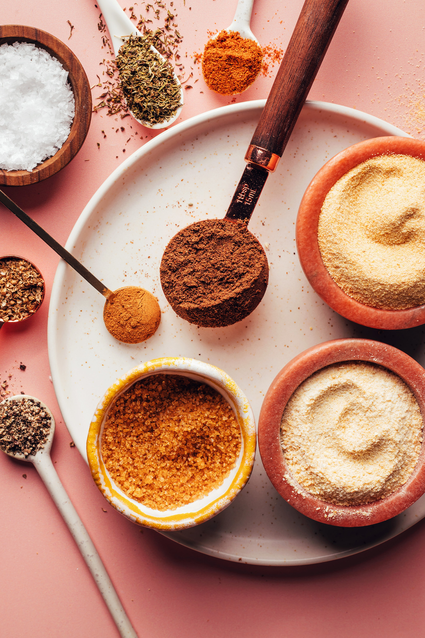 Spoons and bowls of onion powder, garlic powder, allspice, cayenne, thyme, sea salt, cinnamon, coriander, black pepper, and brown sugar