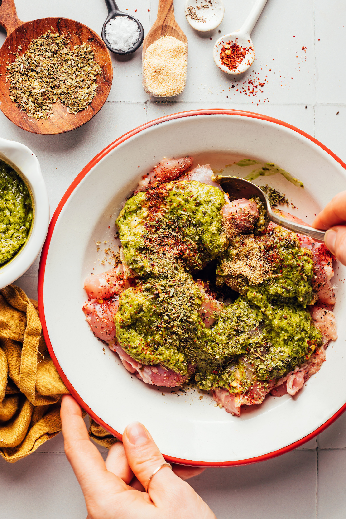 Mixing pesto, dried Italian herbs, red pepper flakes, and chicken thighs in a bowl