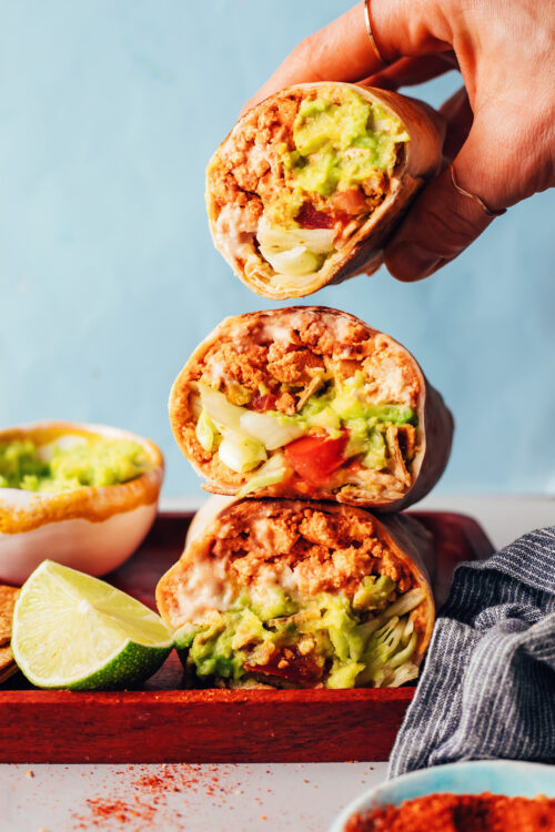 Holding a vegan crunchwrap-inspired burrito above a stack of more burritos