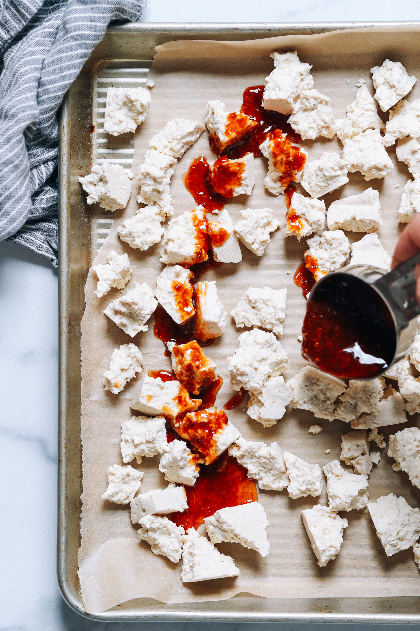 Drizzling a gingery sauce over crumbled tofu on a baking sheet