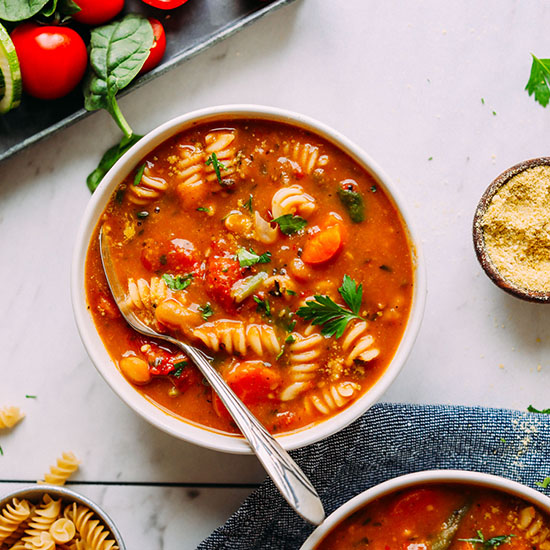 Bowl of Vegan Minestrone beside vegetables, vegan parmesan, and gluten-free noodles