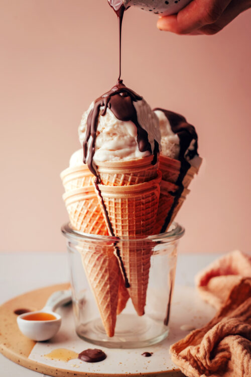 Drizzling chocolate magic shell onto ice cream cones filled with scoops of vegan vanilla ice cream